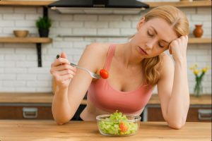 Umorna žena u sportskom topu sjedi za kuhinjskim stolom i nezadovoljno jede salatu iz staklene posude.
