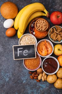 Namirnice bogate željezom s pločicom na kojoj piše Fe, okružene bananama, jajima, žitaricama, mahunarkama, škampima, jetrom, orašastim plodovima i krumpirom.